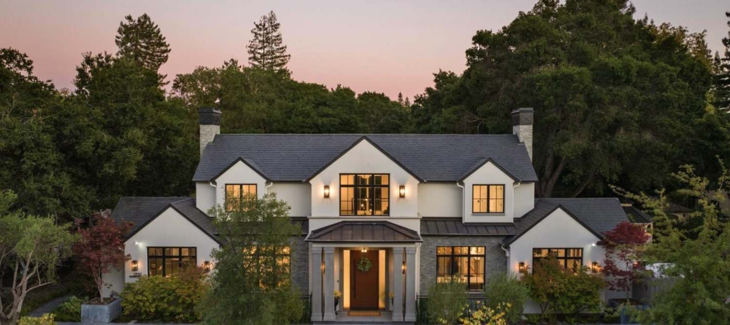 Elegant two-story stucco home with glowing windows at dusk, framed by mature trees