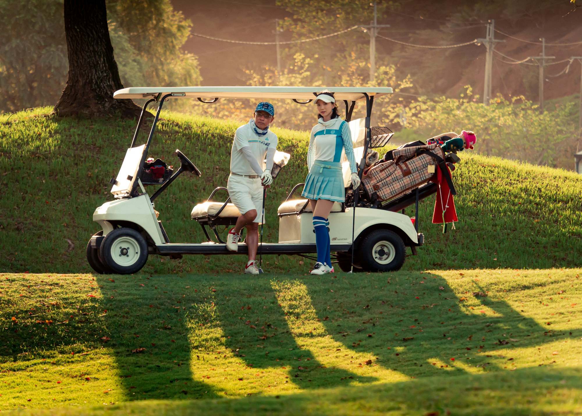Asian couple in golf attire standing by a golf cart on a sunlit course