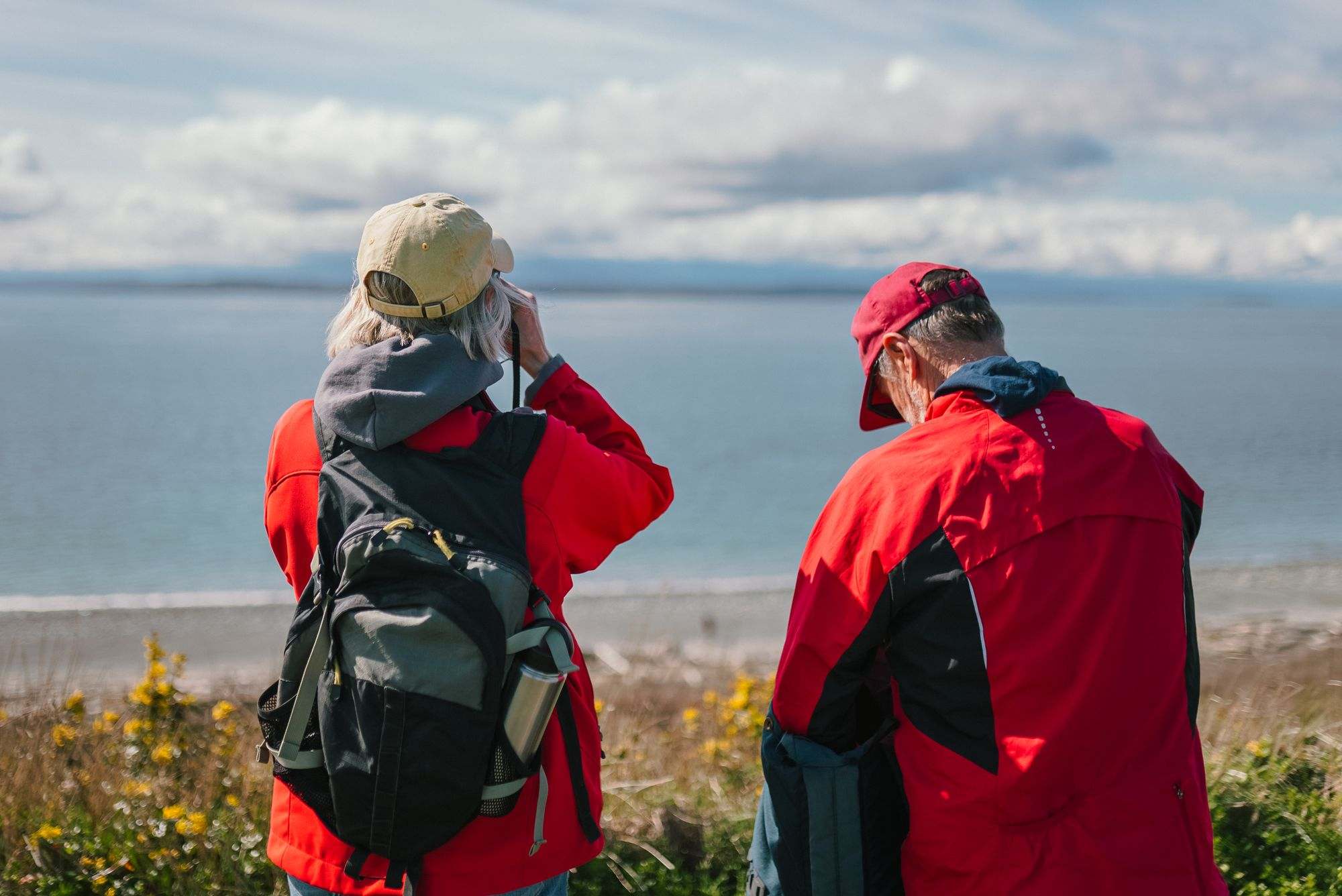 Retired couple in red jackets birdwatching with binoculars on a bluff above the sea